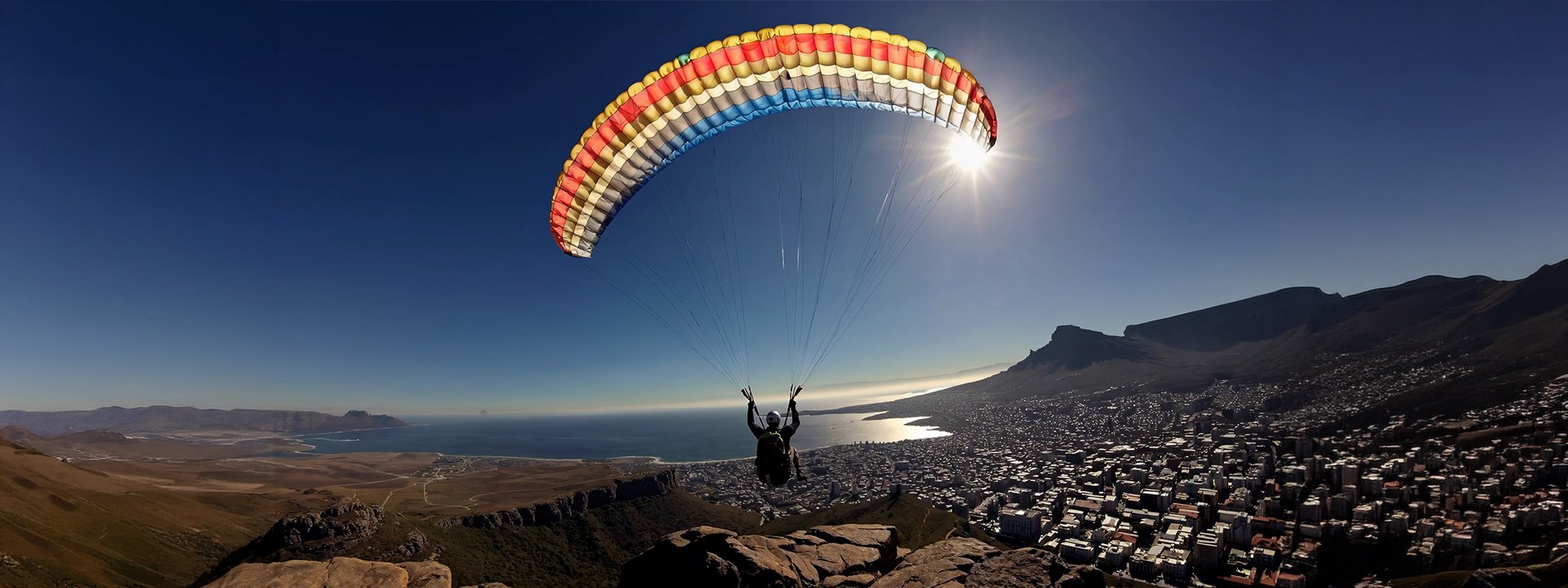 Person paragliding over coastal city landscape with ocean and mountains in the background.