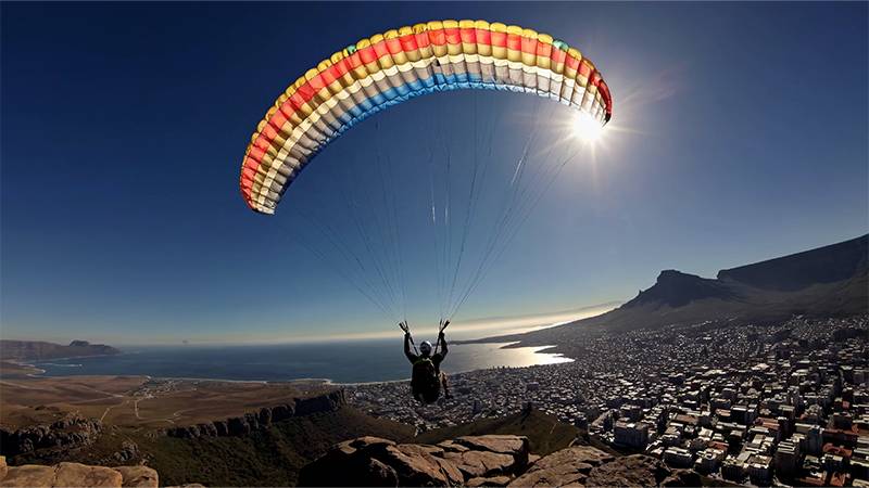 Person paragliding over coastal city landscape with ocean and mountains in the background.