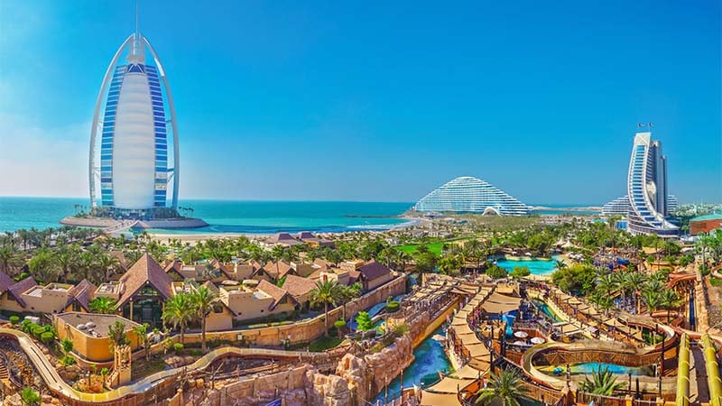 Panoramic view of Dubai with the Burj Al Arab hotel, modern buildings, beaches, and water park attractions under a clear blue sky.
